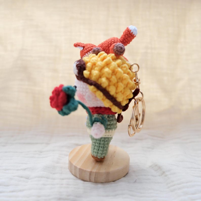 May include: A crochet keychain featuring a small figure with yellow curly hair, a red scarf, and green pants, holding a red flower. An orange fox sits atop the figure's head. The keychain is attached to a gold-toned metal ring and stands on a round wooden base.