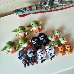 May include: A collection of small, handmade crochet figures featuring green creatures with large ears in orange robes, black-masked figures, white-helmeted troopers, and brown textured characters, arranged alongside a gray and red toy battleship.