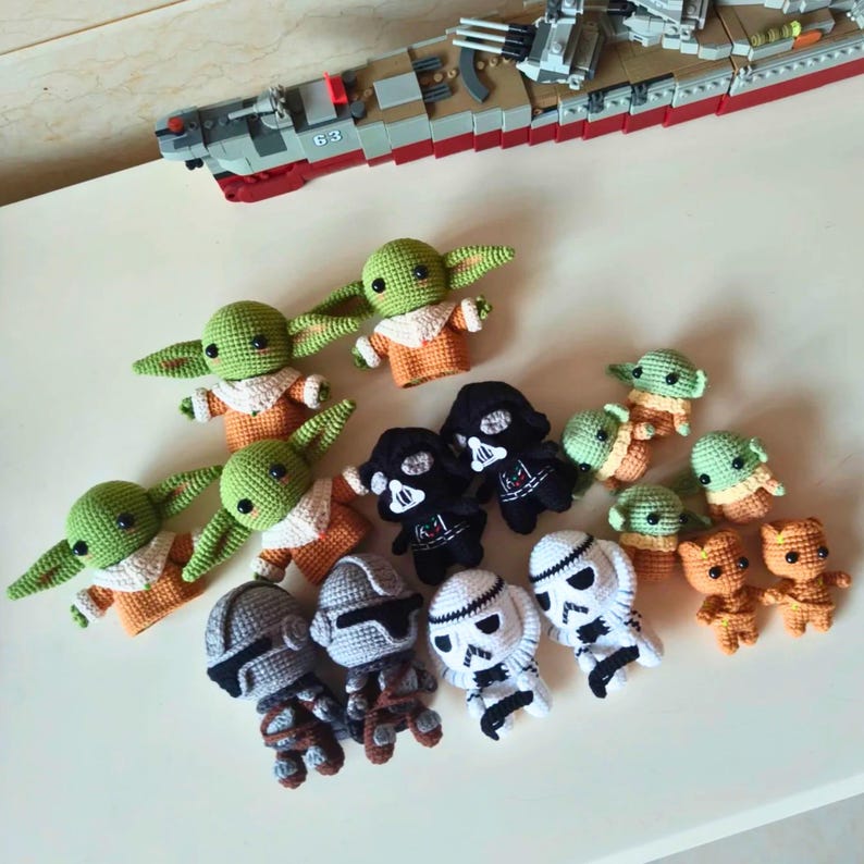 May include: A collection of small, handmade crochet figures featuring green creatures with large ears in orange robes, black-masked figures, white-helmeted troopers, and brown textured characters, arranged alongside a gray and red toy battleship.