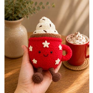 May include: A red crocheted mug plushie featuring a smiling face, white star appliques, and a textured white whipped cream topping with brown flecks. The plush toy has two small brown dangling legs with round feet and a matching red handle.