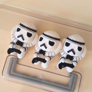 May include: Three handmade crochet dolls designed as stormtroopers. Each doll features a white rounded head with black stitched eye details, a black mouth area, and a white body with black accents. The dolls are arranged in a row, showcasing intricate yarn work and a consistent character design across all three figures.