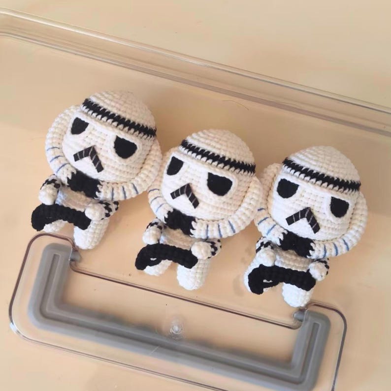 May include: Three handmade crochet dolls designed as stormtroopers. Each doll features a white rounded head with black stitched eye details, a black mouth area, and a white body with black accents. The dolls are arranged in a row, showcasing intricate yarn work and a consistent character design across all three figures.