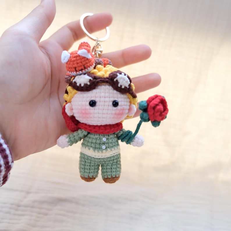 May include: A crocheted keychain featuring a small doll with yellow hair, brown goggles, a red scarf, and a green outfit. The doll holds a red rose in one hand and has a tiny orange and white fox figure perched on top of its head, attached to a metal ring.