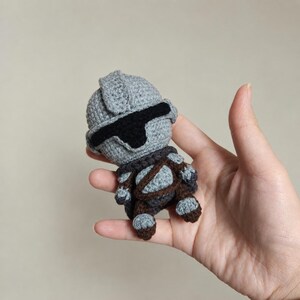 May include: A small, handcrafted crochet doll featuring a gray helmet with a black visor, a dark brown chest piece, and matching brown limbs. The figure is constructed with detailed yarn stitching, showcasing a textured, soft appearance throughout the entire body.