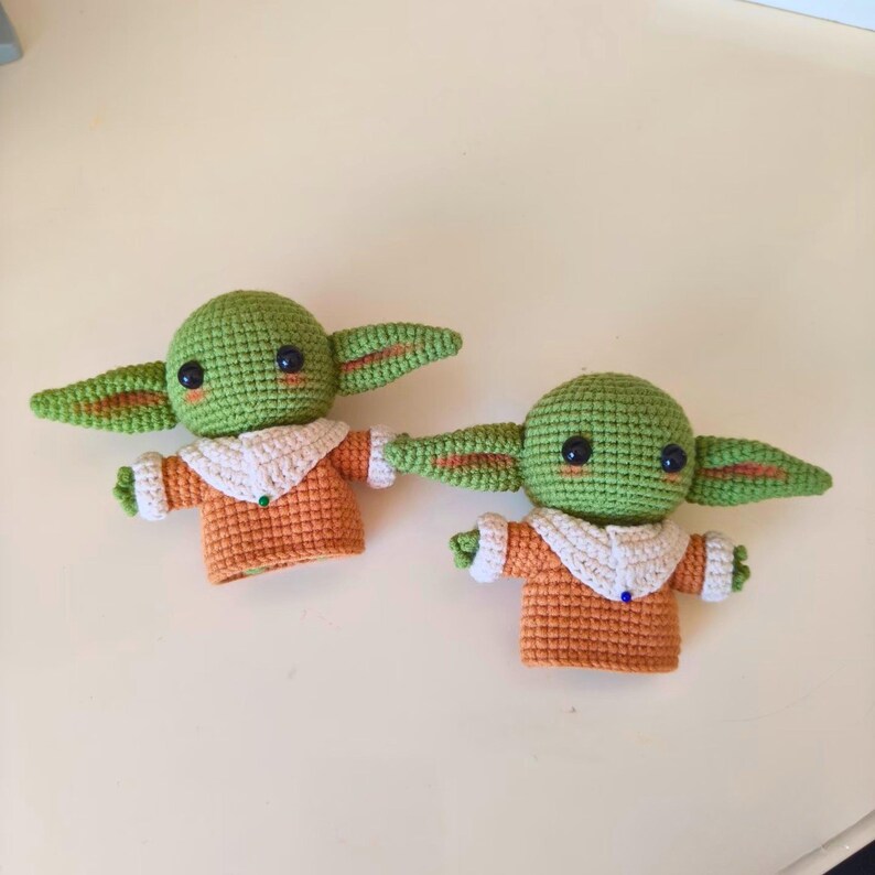 May include: Two handmade crochet dolls featuring green heads with large dark eyes, pointed ears, and pink cheek details. Each doll wears an orange tunic with a white collar and white cuffs, accented by a small blue or green bead at the neckline.