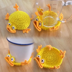 Peut inclure: Un dessous de verre en crochet jaune en forme de canard avec des pattes palmées et un bec orange. Le canard a des yeux blancs avec des pupilles en croix noires. Il est présenté seul, sous une tasse blanche et sous un verre doseur gradué.