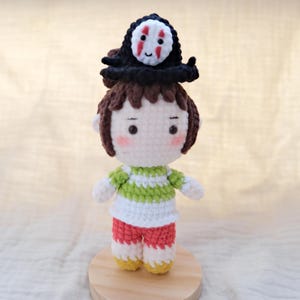 May include: A handcrafted crochet doll featuring brown hair and a black hat topped with a white, red, and black mask-like face. The doll wears a green and white striped shirt, red pants, and yellow shoes, with pink blush on its white yarn face.