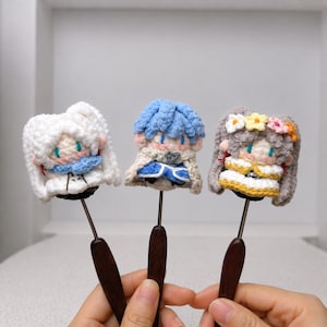 May include: Three crochet doll toppers attached to dark wooden handles. The left doll has white hair and a blue scarf. The center doll has blue hair and a beige cape. The right doll has grey hair with a yellow and pink flower crown and a yellow dress.