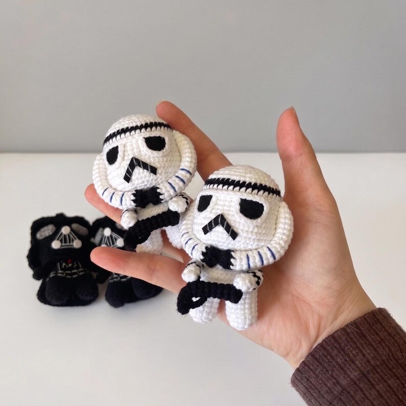 May include: Two small white and black crocheted stormtrooper figures with rounded helmets and black detailing, held in a palm. In the background, two additional black crocheted figures with white accents are visible, featuring detailed chest plates.