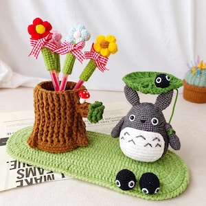 Peut inclure: Un ensemble de bureau en crochet comprenant un porte-stylo en forme de souche d'arbre marron avec trois stylos à fleurs, une figurine grise tenant un parapluie feuille vert et deux petits esprits noirs sur une base ovale verte avec un champignon.