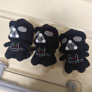 May include: Three black crocheted figures resembling a dark armored character. Each figure features a black helmet, gray eye lenses, a white triangular mouth grill, and a chest panel with small red and green stitched accents on a black body.