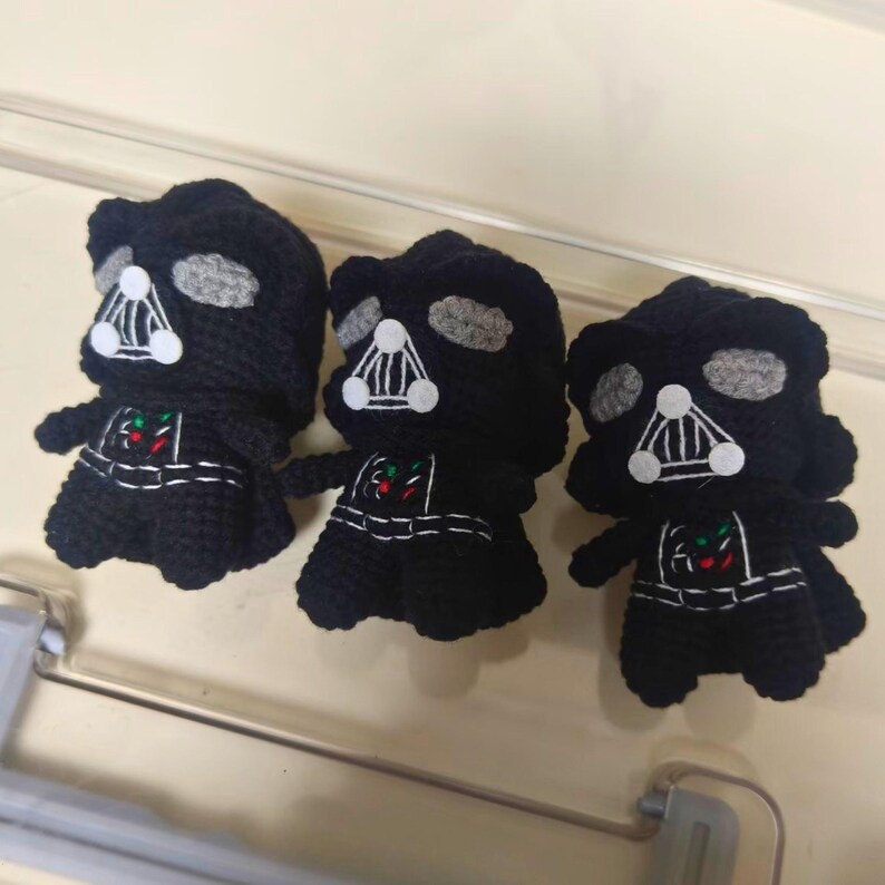 May include: Three black crocheted figures resembling a dark armored character. Each figure features a black helmet, gray eye lenses, a white triangular mouth grill, and a chest panel with small red and green stitched accents on a black body.