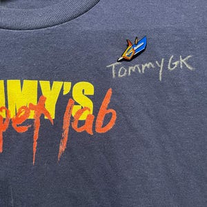 May include: Dark blue t-shirt featuring the text "Tommy's Puppet Lab" in yellow and red. A small pin with a blue and gold design is attached to the shirt, with the handwritten text "TommyGK" next to it.