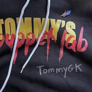 May include: Black hoodie featuring the text "Tommy's Puppet Lab" in yellow and red, with the signature "TommyGK" in white. A small blue and orange pin is attached. White drawstring is visible.
