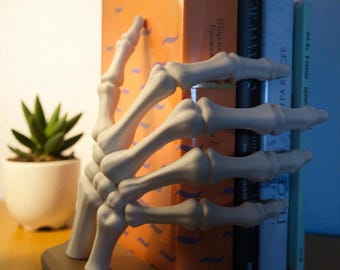 May include: A white skeleton hand bookend holds a stack of books. The bookend is on a brown base, and a small potted succulent is to the left. The books are various colors, including orange, black, and blue.