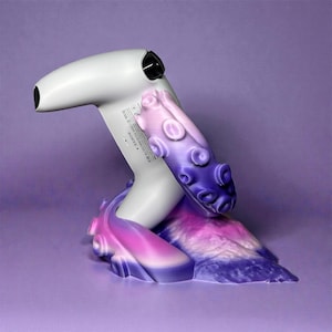 Puede incluir: Un mando de juego blanco es sostenido por un soporte con forma de pulpo morado y rosa. El soporte tiene un esquema de color degradado, que transiciona del rosa al morado. El mando está en ángulo, con su boquilla negra mirando hacia la izquierda.