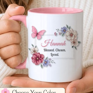 Puede incluir: Una taza de cerámica blanca con interior y asa de color rosa. El exterior presenta un marco rectangular con el nombre Hannah y el texto Blessed. Chosen. Loved. El diseño está decorado con flores rosas y moradas y una mariposa rosa.