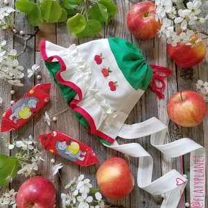 May include: A white and green fabric baby bonnet with red trim and embroidered apples. The bonnet is tied with a white ribbon and is surrounded by white blossoms, red apples, and wrapped candies on a wooden surface.