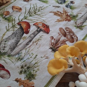 May include: A white tea towel with a printed design of mushrooms and leaves. The mushrooms are in various shades of brown, red, and yellow. The leaves are green and brown. The tea towel is folded in half and has a green border.