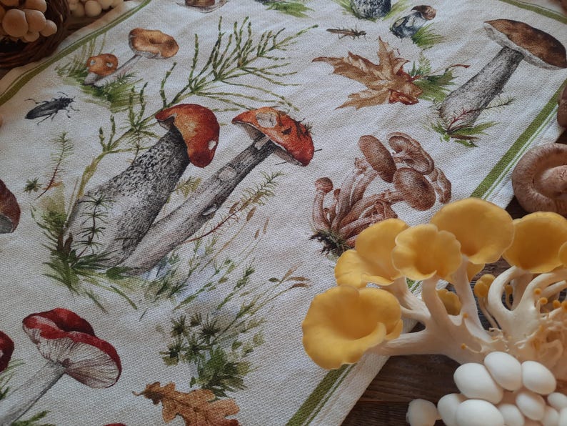 May include: A white tea towel with a printed design of mushrooms and leaves. The mushrooms are in various shades of brown, red, and yellow. The leaves are green and brown. The tea towel is folded in half and has a green border.
