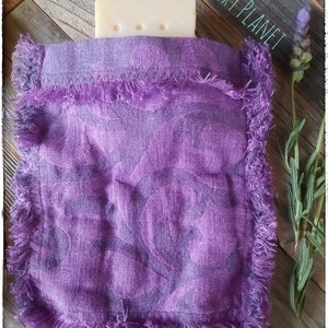 Puede incluir: Una bolsa morada para jab&oacute;n con un borde con flecos contiene una pastilla de jab&oacute;n. La bolsa tiene un aspecto texturizado y est&aacute; hecha de un material tejido. Una ramita de lavanda se coloca a la derecha de la bolsa.