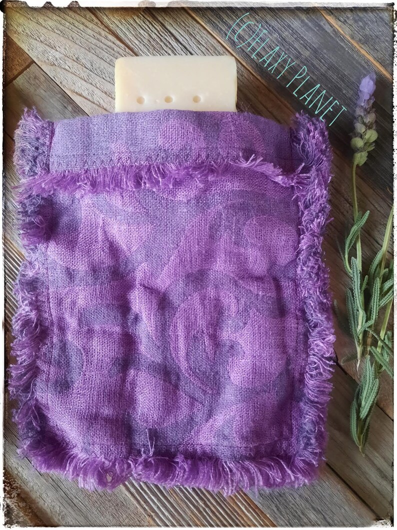 Puede incluir: Una bolsa morada para jab&oacute;n con un borde con flecos contiene una pastilla de jab&oacute;n. La bolsa tiene un aspecto texturizado y est&aacute; hecha de un material tejido. Una ramita de lavanda se coloca a la derecha de la bolsa.