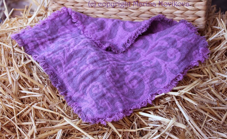 Puede incluir: Un pa&ntilde;uelo cuadrado de lino morado con un estampado floral y bordes deshilachados. El pa&ntilde;uelo se muestra sobre un lecho de paja, con una cesta de mimbre en el fondo. Las dimensiones del pa&ntilde;uelo no son visibles.