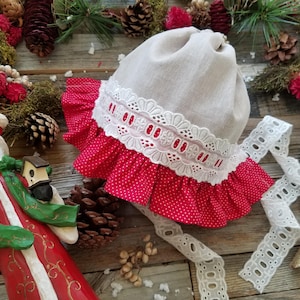 May include: A white linen baby bonnet with a red and white polka dot ruffle and white lace trim. The bonnet is lying on a wooden surface with Christmas decorations.