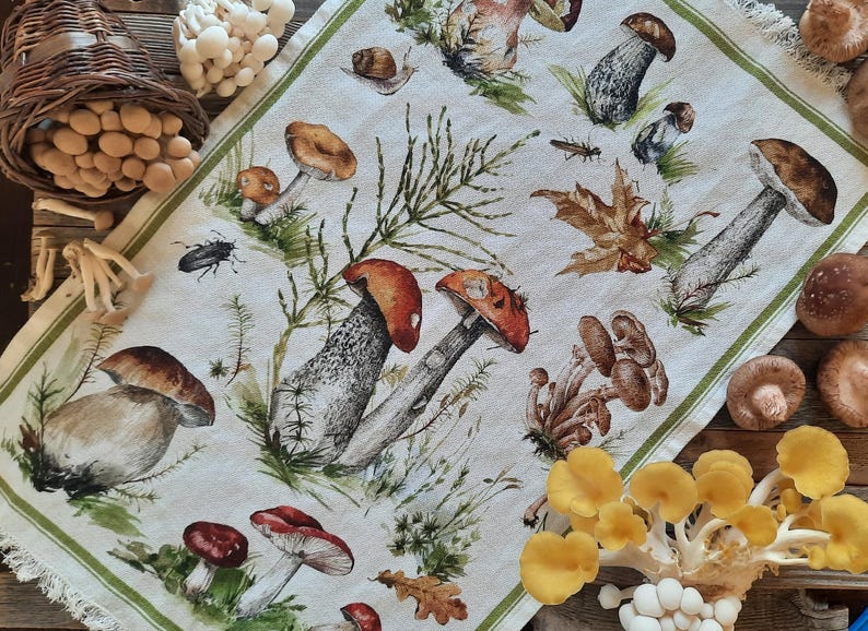 May include: A white kitchen towel with a green border features a watercolor illustration of various mushrooms in shades of brown, red, and yellow. The mushrooms are depicted in different sizes and shapes, with some growing in clusters and others standing alone. The towel is a decorative kitchen accessory.