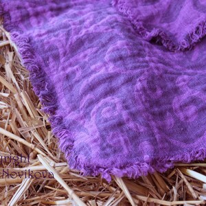 Puede incluir: Primer plano de un textil de lino morado con un estampado floral y bordes deshilachados. La tela est&aacute; drapeada sobre paja seca, creando un contraste r&uacute;stico. El color del textil es un tono vibrante de morado.