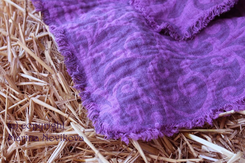 Puede incluir: Primer plano de un textil de lino morado con un estampado floral y bordes deshilachados. La tela est&aacute; drapeada sobre paja seca, creando un contraste r&uacute;stico. El color del textil es un tono vibrante de morado.