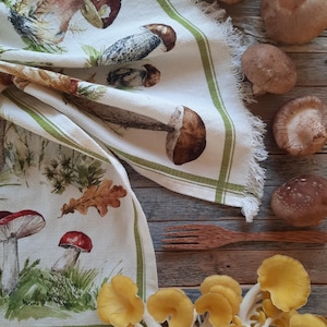 May include: A white tea towel with a green border and a mushroom print. The towel is folded and lying on a wooden surface with various mushrooms scattered around it. The mushrooms are a mix of brown, white, and yellow.