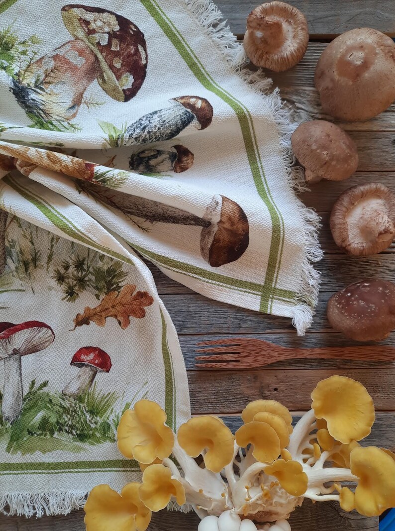 May include: A white tea towel with a green border and a mushroom print. The towel is folded and lying on a wooden surface with various mushrooms scattered around it. The mushrooms are a mix of brown, white, and yellow.