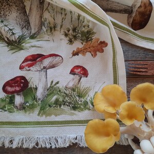 May include: A white tea towel with a green border and a fringe hem. The tea towel features a watercolor illustration of mushrooms and leaves.