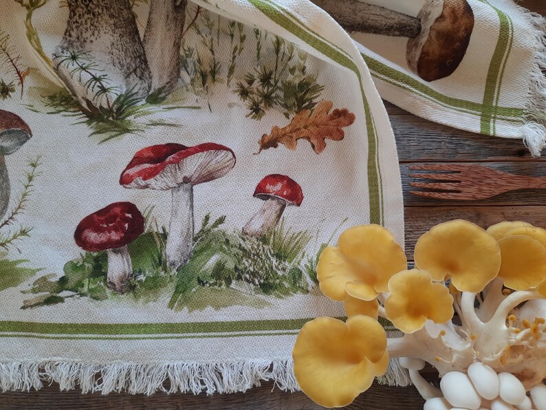 May include: A white tea towel with a green border and a fringe hem. The tea towel features a watercolor illustration of mushrooms and leaves.
