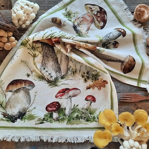 May include: A white tea towel with a green border features a watercolor illustration of various mushrooms in shades of brown, red, and white. The mushrooms are depicted in a realistic style with intricate details.