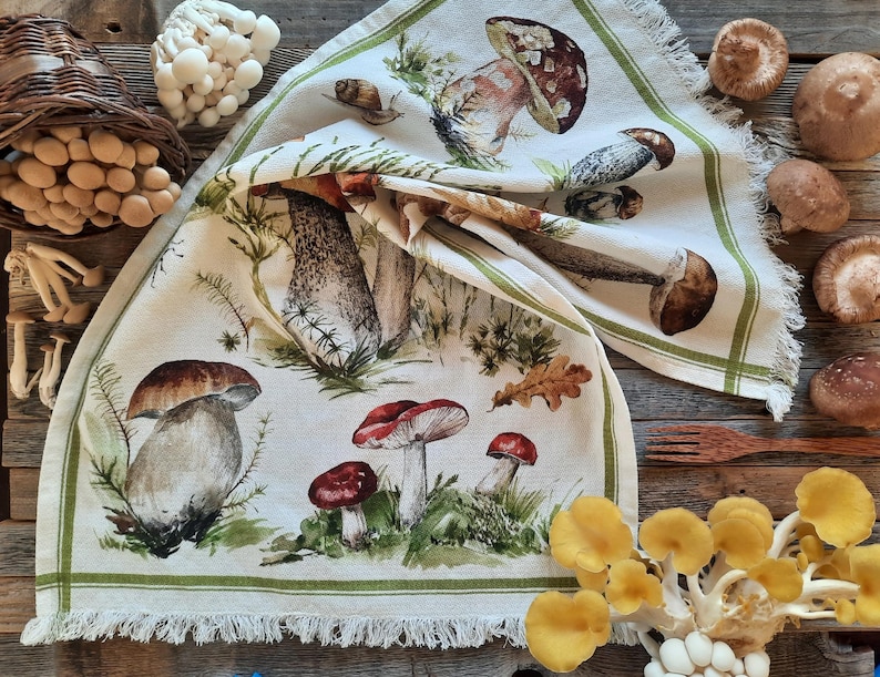 May include: A white tea towel with a green border features a watercolor illustration of various mushrooms in shades of brown, red, and white. The mushrooms are depicted in a realistic style with intricate details.