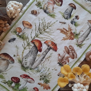 May include: A white tea towel with a green border features a colorful illustration of various mushrooms and toadstools. The design includes red, brown, white, and yellow mushrooms with green leaves and stems.