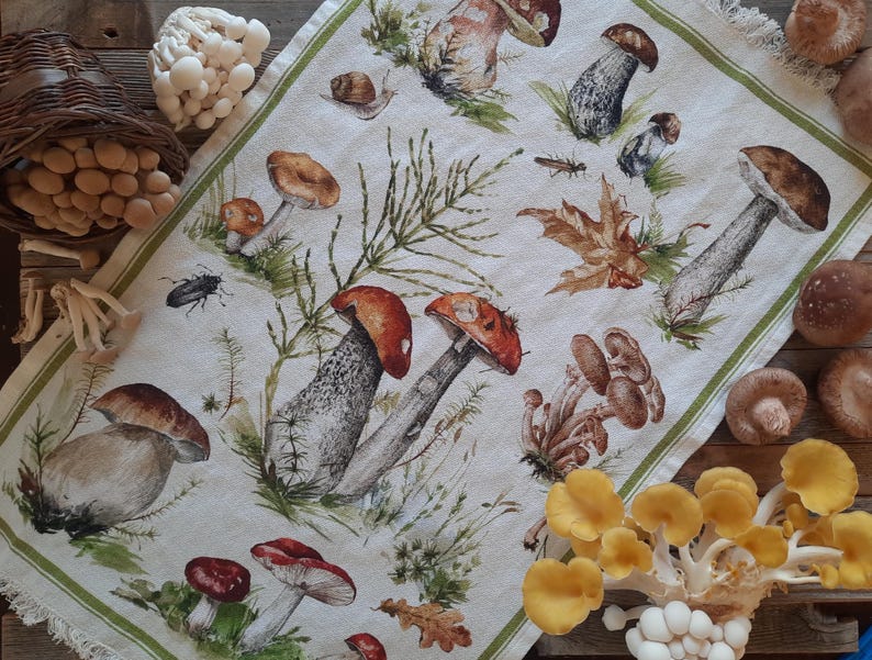 May include: A white tea towel with a green border features a colorful illustration of various mushrooms and toadstools. The design includes red, brown, white, and yellow mushrooms with green leaves and stems.