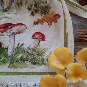 May include: A white tea towel with a green border and a fringe. The towel features a watercolor illustration of mushrooms, leaves, and grass.