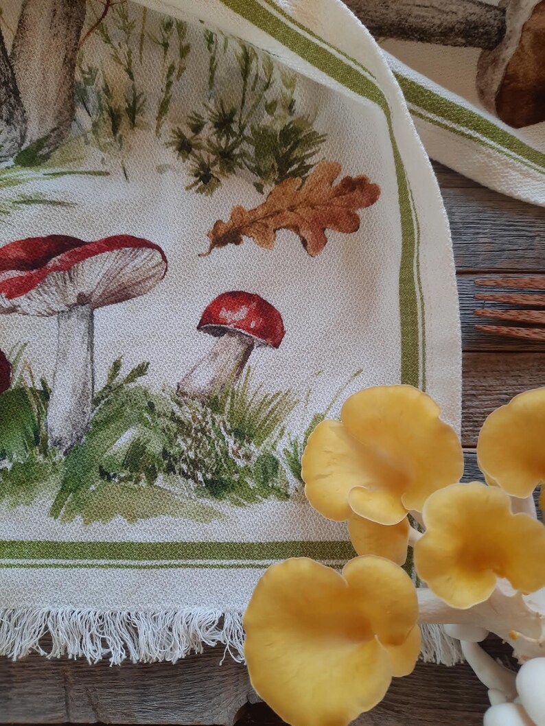 May include: A white tea towel with a green border and a fringe. The towel features a watercolor illustration of mushrooms, leaves, and grass.