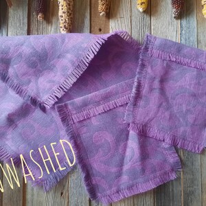 Puede incluir: Un juego de servilletas de lino moradas y un camino de mesa a juego con un borde con flecos. Las servilletas y el camino de mesa tienen un patr&oacute;n de remolino morado m&aacute;s oscuro. La palabra "UNWASHED" est&aacute; escrita en amarillo claro.
