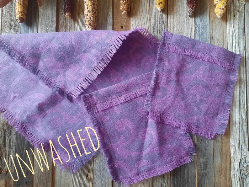Puede incluir: Un juego de servilletas de lino moradas y un camino de mesa a juego con un borde con flecos. Las servilletas y el camino de mesa tienen un patr&oacute;n de remolino morado m&aacute;s oscuro. La palabra "UNWASHED" est&aacute; escrita en amarillo claro.