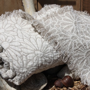May include: Two beige linen throws with a white floral pattern and frayed edges. The throws are folded and stacked on top of each other.