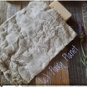 May include: A white bar of soap wrapped in a natural linen fabric pouch with frayed edges. The pouch is on a wooden surface with lavender sprigs.
