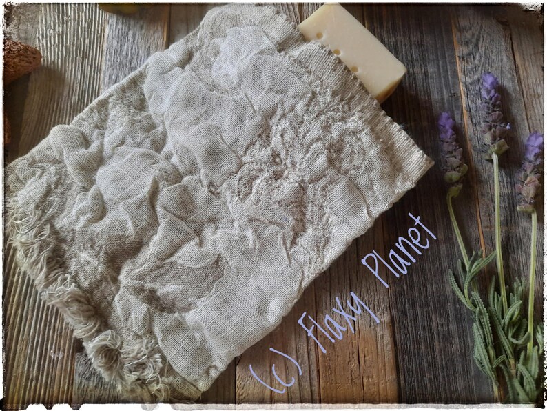 May include: A white bar of soap wrapped in a natural linen fabric pouch with frayed edges. The pouch is on a wooden surface with lavender sprigs.