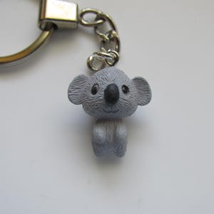 May include: A silver-toned metal keyring featuring a small, textured grey koala figurine charm. The koala has a round head, large ears, a prominent black nose, and small black eyes, suspended from a standard circular split ring by a short chain.