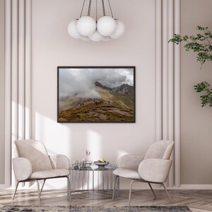 May include: A framed landscape photograph of a mountain range shrouded in mist. The image features a dark frame, with a view of the mountains and a cloudy sky. The photograph is displayed above two beige armchairs and a small glass table.