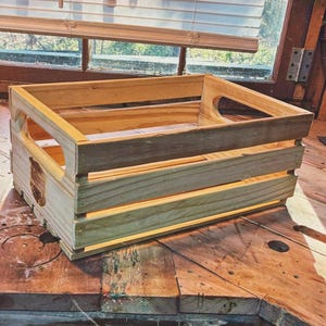May include: A handcrafted wooden crate with a rectangular shape and open sides. The crate features a natural wood finish with visible grain and a handle cutout on each end. The crate is sitting on a wooden surface.