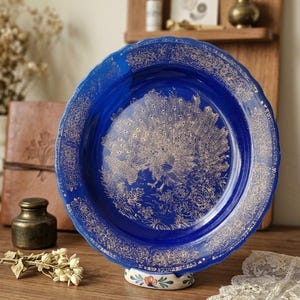 Puede incluir: Un plato decorativo azul vibrante con un diseño de pavo real dorado en el centro. El plato tiene un borde festoneado y detalles dorados intrincados alrededor del borde. Descansa sobre una base blanca con detalles florales.